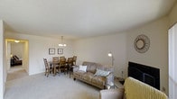 This single-floor space presents an open-concept living and dining area unified by a neutral color palette and soft, plush carpeting throughout. The living area is anchored by a comfortable upholstered sofa accented with decorative pillows, positioned alongside a floor lamp and television, creating a cozy and functional lounge space. A textured ceiling adds subtle visual depth overhead.

Adjacent to the living area, the dining space accommodates a full wooden table with seating for six, making it well-suited for gatherings and everyday meals. A classic chandelier with multiple pendant lights hangs above the dining table, providing warm, focused illumination. Two framed artworks are displayed on the wall, contributing to a clean, understated aesthetic.

The open layout between the living and dining zones allows for natural flow and versatile use of the space. Warm beige walls complement the light carpeting, reinforcing the inviting and calm ambiance throughout. The overall design is practical yet comfortable, with a straightforward arrangement that maximizes usability across the shared living spaces.