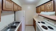 This single-floor unit is designed with practicality and comfort in mind, offering a well-organized layout across its key living spaces. The kitchen is a galley-style space lined with warm oak cabinetry on both sides, providing ample storage. White appliances — including a coil-burner electric range and a full-sized refrigerator — complement the light countertops, while a double basin sink adds convenience. Light tile flooring runs throughout, creating a cohesive and easy-to-maintain surface across the unit. The living and bedroom areas are bright and open, featuring cream-toned walls, textured ceilings, and windows that allow natural light to filter in while offering views of a verdant outdoor landscape. Wood-trimmed baseboards add a subtle warmth to these otherwise clean and neutral spaces. Each room includes closet access with wood-framed sliding doors. The bathroom is cleanly finished, featuring a tiled tub-and-shower combination with a chrome curtain rod, a wall-mounted vanity sink, and a wood-framed medicine cabinet with vanity lighting above. The overall ambiance of the unit is understated and functional, presenting a blank canvas that is move-in ready and adaptable to a variety of living arrangements.