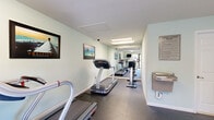 This dedicated cardio studio fitness center offers a well-organized and functional workout space designed to support cardiovascular exercise. The room features durable rubber flooring throughout, providing a safe and slip-resistant surface suited for high-traffic fitness use. The light mint-green walls contribute to a clean, refreshing ambiance that keeps the space feeling open and energizing despite its compact footprint.

The facility is equipped with multiple treadmills, including Star Trac models, as well as elliptical cross-trainers, offering users a solid variety of cardio options. A wall-mounted flat-screen television provides entertainment during workouts. A built-in water fountain is conveniently installed along one wall, and posted cardio studio rules ensure the space is used appropriately by all guests.

Natural light filters in through windows fitted with blinds, and a glass entry door opens to an outdoor walkway, further brightening the interior. Overhead fluorescent and recessed lighting ensures the space remains well-lit at all times. Motivational framed artwork — themed around ambition, destiny, and vision — is displayed throughout the room, adding an encouraging atmosphere. A wall-mounted air conditioning unit provides climate control for year-round comfort. The studio is a purpose-built, efficiently utilized fitness amenity.
