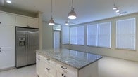 This single-floor space offers a clean, modern aesthetic centered around an open-concept layout that seamlessly connects the kitchen and dining areas. The kitchen is a standout feature, anchored by a generous island topped with polished granite countertops featuring subtle veining that adds both elegance and visual interest. White shaker-style cabinetry with dark hardware lines the kitchen, complementing the stainless steel refrigerator and maintaining a crisp, cohesive look throughout. Two industrial-style chrome pendant lights hang above the island, providing focused task lighting while contributing to the space's contemporary character. The adjacent dining area is bathed in natural light, courtesy of a wide bank of windows dressed with white horizontal blinds that allow for flexible light control. A ceiling fan positioned centrally in this area ensures comfort and airflow throughout. The overall color palette — white cabinetry, light walls, and bright natural light — creates an airy, welcoming atmosphere. The open flow between functional zones makes the space feel expansive and well-suited for both everyday living and entertaining.