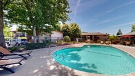 Located at 5101 Hackberry Ln in Sacramento, California, this single-level outdoor community space centers around a generously sized swimming pool with clear, blue-green water. The pool features a classic curved design with clearly marked depth indicators and is accessible via built-in entry steps, making it welcoming for a range of swimmers.

Surrounding the pool is a spacious concrete deck that provides ample room for lounging and movement. Outdoor seating is available poolside, including lounge chairs and a shaded dining area with a table, chairs, and a bold red umbrella — ideal for relaxing in the Sacramento sun. Safety equipment, including a life ring, is mounted conveniently along the perimeter fencing.

The area is enclosed by a sturdy wrought iron fence that provides security while maintaining an open, airy feel. Towering mature trees cast natural shade across much of the deck, creating a cool and pleasant atmosphere during warmer months. Well-maintained landscaping with shrubs and ground cover borders the fence line, adding a touch of greenery to the space.

An adjacent single-story structure, likely a clubhouse or community facility, is visible at the edge of the pool area, complementing the overall layout. The combination of thoughtful amenities, natural shade, and carefully maintained surroundings makes this outdoor space a comfortable and functional community gathering area.