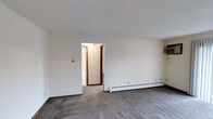 This single-floor unit offers a functional layout within a compact but efficiently organized footprint. The living area features patterned wall-to-wall carpeting that adds texture underfoot, while clean white walls and a smooth white ceiling create a bright, neutral backdrop throughout the space. A baseboard heating unit runs along one wall of the living area, providing an efficient and consistent heat source. Dark wood-toned baseboards and door trim provide subtle contrast against the light walls, lending a modest architectural detail to the interior. An open doorway connects the living area to the remainder of the unit, which includes one bedroom and one full bathroom. The layout appears straightforward and easy to navigate, with a focus on practical use of the available space. The overall ambiance is clean and understated, offering a blank-canvas interior well suited for personalizing with furnishings and décor. The unit is designed to deliver essential, comfortable living in a manageable, easy-to-maintain space.
