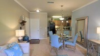 Located at 8800 Pine Forest Rd in Pensacola, Florida, this single-floor residence offers a thoughtfully designed open-concept layout that seamlessly connects the living, dining, and kitchen areas. The interior is finished with warm, neutral tones throughout, creating a cohesive and inviting atmosphere. Crown molding adds a refined architectural detail along the ceilings, lending a polished character to the main living space. The dining area is highlighted by an elegant pendant light fixture that serves as a focal point, complementing the glass-top dining table positioned at the heart of the room. Plush carpeting underfoot adds warmth and comfort to the living and dining zones. The kitchen is efficiently designed with warm wood-toned cabinetry, ample counter space, and full-sized appliances, making it both functional and visually appealing. A louvered-door closet near the entryway provides convenient storage. Natural and ambient lighting work together to keep the space feeling bright and welcoming. The overall layout and design offer a comfortable, well-appointed living environment well-suited to everyday living in the Pensacola area.