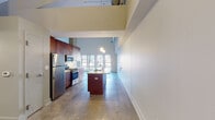 This single-floor loft-style unit showcases an industrial-modern aesthetic defined by dramatically high ceilings with exposed ductwork and a polished concrete floor that flows throughout the main living and kitchen areas. The open-plan living space is anchored by a wall of large windows fitted with an ornate railing, framing striking urban skyline views and flooding the interior with natural light.

The kitchen is thoughtfully designed with rich warm-toned wood cabinetry, light solid-surface countertops, stainless steel appliances including a gas range, microwave, and refrigerator, and a central island with a sink. A raised platform subtly delineates the kitchen zone from the main living area. A stylish barn-style sliding door with frosted glass panels provides access to an adjoining room, adding a contemporary architectural detail.

The two bedrooms are carpeted and offer a quieter, softer contrast to the polished concrete elsewhere. Each is accessed through private doorways and provides ample wall space. The unit includes two bathrooms, both featuring warm-toned tile flooring, wood vanities with light countertops, and pendant lighting. An in-unit washer and dryer are also present. The overall ambiance is open, urban, and loft-inspired.