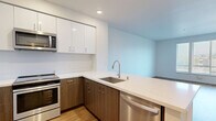 Located at 3301 E 12th Street in Oakland, CA, this modern one-bedroom, one-bathroom unit offers a well-designed layout across a single floor. The kitchen is a standout feature, showcasing two-tone cabinetry with sleek white upper cabinets and warm wood-toned lower cabinets, complemented by quartz countertops, a stainless steel undermount sink, and a full suite of stainless steel appliances including a refrigerator, electric range, built-in microwave, and dishwasher. Light wood-style flooring runs throughout the main living areas, contributing to a bright and cohesive aesthetic. The open kitchen flows naturally into the living area, which benefits from large windows fitted with blinds that invite natural light while providing views of the surrounding neighborhood. The bedroom features plush carpeting, a sliding-door closet with built-in shelving and a hanging rod, and in-unit washer and dryer hookups. The bathroom is finished with wood-style flooring, a full tub-shower combo, and clean white wall surrounds. Recessed lighting and crisp white walls throughout the unit create a clean, contemporary ambiance well-suited for modern urban living.