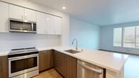 Located at 3301 E 12th Street in Oakland, CA, this modern one-bedroom, one-bathroom unit offers a well-designed layout across a single floor. The kitchen is a standout feature, showcasing two-tone cabinetry with sleek white upper cabinets and warm wood-toned lower cabinets, complemented by quartz countertops, a stainless steel undermount sink, and a full suite of stainless steel appliances including a refrigerator, electric range, built-in microwave, and dishwasher. Light wood-style flooring runs throughout the main living areas, contributing to a bright and cohesive aesthetic. The open kitchen flows naturally into the living area, which benefits from large windows fitted with blinds that invite natural light while providing views of the surrounding neighborhood. The bedroom features plush carpeting, a sliding-door closet with built-in shelving and a hanging rod, and in-unit washer and dryer hookups. The bathroom is finished with wood-style flooring, a full tub-shower combo, and clean white wall surrounds. Recessed lighting and crisp white walls throughout the unit create a clean, contemporary ambiance well-suited for modern urban living.