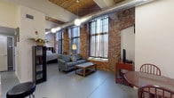 This furnished studio showcases an industrial loft aesthetic rooted in authentic architectural details. The main living area is defined by striking exposed brick columns and walls, soaring ceilings with warm wood paneling, and exposed ductwork that adds to the space's character. Tall, grid-paned windows dressed with sheer curtains flood the room with natural light and feature built-in window seats. The open layout accommodates a living area with a sofa, coffee table, and television on a red media console, alongside a sleeping area separated by a bookshelf divider.

The fully equipped kitchen is a standout feature, finished with dark espresso cabinetry extending to the ceiling, granite countertops, and a central island with bar seating. Black appliances, including a refrigerator, oven, and dishwasher, complement the bold cabinetry. Pendant lighting illuminates both the island and the surrounding workspace.

The bathroom is cleanly finished with slate tile flooring, a floating dark vanity with a large mirror, and accessibility features including grab bars and a raised toilet. Throughout, polished concrete floors tie the industrial and contemporary elements together cohesively.