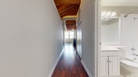 This single-floor loft-style unit showcases a compelling blend of industrial character and contemporary design. The standout architectural feature is the soaring exposed wood beam and tongue-and-groove ceiling, which spans the main living area and hallway, lending warmth and a distinctly warehouse-inspired aesthetic. Visible ductwork and track lighting complement the raw, open overhead structure.

Rich, dark hardwood flooring runs throughout the main living spaces and hallway, reflecting light and adding a polished contrast to the rustic ceiling above. The open-concept living area flows seamlessly into the kitchen, which is equipped with warm cherry cabinetry, dark granite countertops, black appliances including a refrigerator, range, and built-in microwave, and a decorative wine rack above the upper cabinets. A partial peninsula helps define the kitchen from the living space.

A long, well-proportioned hallway connects the main area to the private rooms. The bedroom features tall ceilings, plush neutral carpeting, and a window with natural light. The bathroom offers a clean, functional design with white cabinetry, tile flooring, and a large vanity mirror. Overall, the space delivers an open, airy industrial ambiance with thoughtful modern finishes throughout.