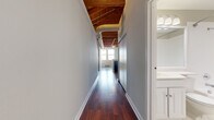 This single-floor loft-style unit showcases a compelling blend of industrial character and contemporary design. The standout architectural feature is the soaring exposed wood beam and tongue-and-groove ceiling, which spans the main living area and hallway, lending warmth and a distinctly warehouse-inspired aesthetic. Visible ductwork and track lighting complement the raw, open overhead structure.

Rich, dark hardwood flooring runs throughout the main living spaces and hallway, reflecting light and adding a polished contrast to the rustic ceiling above. The open-concept living area flows seamlessly into the kitchen, which is equipped with warm cherry cabinetry, dark granite countertops, black appliances including a refrigerator, range, and built-in microwave, and a decorative wine rack above the upper cabinets. A partial peninsula helps define the kitchen from the living space.

A long, well-proportioned hallway connects the main area to the private rooms. The bedroom features tall ceilings, plush neutral carpeting, and a window with natural light. The bathroom offers a clean, functional design with white cabinetry, tile flooring, and a large vanity mirror. Overall, the space delivers an open, airy industrial ambiance with thoughtful modern finishes throughout.