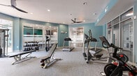 This fitness center is a thoughtfully designed workout space that offers a comprehensive range of equipment within a compact yet efficiently utilized floor plan. The interior is finished with teal-blue accent walls and a neutral white ceiling, creating a clean and energizing atmosphere. Recessed lighting and a ceiling fan ensure consistent illumination and air circulation throughout the space.

The cardio section features multiple True-brand treadmills and at least one elliptical trainer, providing ample options for aerobic exercise. A dedicated free weight area includes a tiered dumbbell rack stocked with a wide range of weights, accompanied by adjustable workout benches for strength training. A cable machine and resistance training equipment round out the strength offerings.

Floor-to-ceiling mirrors line one wall, visually expanding the space while allowing users to monitor their form during exercise. A wall-mounted flat-screen TV adds an entertainment element to cardio workouts. Large floor-to-ceiling windows along one side of the room invite natural light and offer views of an outdoor area, contributing to an open and motivating environment. Frosted glass panel partitions provide visual separation from adjacent spaces while maintaining an airy feel. The carpeted flooring adds comfort underfoot throughout the training area.