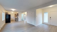 This single-floor unit is designed with an open-concept layout that seamlessly connects the living area and kitchen, creating a functional and cohesive living environment. Light hardwood-style flooring runs throughout the space, contributing to a clean, contemporary aesthetic.

The kitchen is partially enclosed by a floor-to-ceiling dark wood-toned cabinetry partition, which adds visual definition between the cooking and living zones while maintaining an open feel. The kitchen features dark lower and upper cabinetry with a contrasting light countertop, a stainless steel sink, and a stainless steel refrigerator. A track lighting fixture mounted to the ceiling provides focused task lighting over the kitchen area.

The living area is generously proportioned, offering ample room for furnishing arrangements. A window with a roller blind allows natural light to filter in while providing privacy. A baseboard heater runs along the exterior wall beneath the window, providing efficient heating. The textured ceiling adds subtle architectural detail throughout. Overall, the space presents a practical, modern interior with a neutral palette suited to a variety of decor styles.