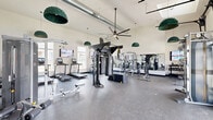Located at 6205 Longstem Way in Loveland, Colorado, this fitness center at Bella Terra offers a generously sized, single-floor exercise space designed to accommodate a full range of workout routines. The room features soaring vaulted ceilings with exposed ductwork and large ceiling fans, creating an open, airy atmosphere that is both functional and inviting.

Natural light pours in through multiple windows and a skylight, brightening the durable rubber-speckled flooring throughout. The space is thoughtfully organized into distinct workout zones. A dedicated cardio area houses multiple treadmills, elliptical machines, and an air bike, each equipped with individual display screens, alongside a rowing machine and wall-mounted TVs for entertainment during workouts.

The strength and functional training zone is anchored by a central multi-purpose cable and functional trainer rack, complemented by TRX suspension trainers, battle ropes, and a plyo box. A full dumbbell rack and selectorized cable machines line the walls, offering options for both free weight and guided resistance training. Floor-to-ceiling mirrors expand the sense of space and allow for form monitoring.

Distinctive green pendant lighting adds a stylish touch, while the Bella Terra logo is boldly displayed on the wall, reinforcing the community identity of this well-appointed facility.