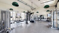 Located at 6205 Longstem Way in Loveland, Colorado, this fitness center at Bella Terra offers a generously sized, single-floor exercise space designed to accommodate a full range of workout routines. The room features soaring vaulted ceilings with exposed ductwork and large ceiling fans, creating an open, airy atmosphere that is both functional and inviting.

Natural light pours in through multiple windows and a skylight, brightening the durable rubber-speckled flooring throughout. The space is thoughtfully organized into distinct workout zones. A dedicated cardio area houses multiple treadmills, elliptical machines, and an air bike, each equipped with individual display screens, alongside a rowing machine and wall-mounted TVs for entertainment during workouts.

The strength and functional training zone is anchored by a central multi-purpose cable and functional trainer rack, complemented by TRX suspension trainers, battle ropes, and a plyo box. A full dumbbell rack and selectorized cable machines line the walls, offering options for both free weight and guided resistance training. Floor-to-ceiling mirrors expand the sense of space and allow for form monitoring.

Distinctive green pendant lighting adds a stylish touch, while the Bella Terra logo is boldly displayed on the wall, reinforcing the community identity of this well-appointed facility.