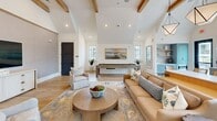 Located at 350 Henslow Dr in Charleston, SC, this single-floor community space offers a welcoming and well-appointed gathering environment. The living room serves as the heart of the space, defined by dramatic vaulted ceilings adorned with exposed wood beams that lend warmth and architectural character. Light wide-plank hardwood flooring runs throughout, complementing the crisp white walls and contributing to a bright, airy coastal aesthetic. Comfortable, curated furnishings — including plush sofas, accent chairs, and a round wood coffee table — create an inviting lounge atmosphere, while a wall-mounted television adds a media viewing option. A billiards table positioned at one end of the room provides an additional recreational feature. The kitchenette area is thoughtfully designed with soft grey cabinetry accented by gold hardware, a white countertop workspace, and a sink beneath an expansive bank of multi-pane windows that flood the area with natural light and offer outdoor views. A large white communal dining table with rattan chairs provides generous seating. Open shelving in soft blue tones adds a charming coastal touch. Colorful landscape artwork and geometric pendant lighting complete the refined, relaxed ambiance of this well-designed community gathering space.