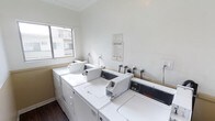 This shared laundry facility is designed for practicality and convenience, offering residents access to multiple commercial-grade washing machines and dryers arranged in a well-organized layout. The room features crisp white walls accented by crown molding near the ceiling, lending a clean and polished appearance to the utilitarian space. Dark hardwood-style flooring contrasts with the bright white appliances and walls, adding a subtle design element to the room. A window on the exterior wall allows natural light to fill the space, creating a bright and pleasant environment. Posted laundry room rules and operating hours are clearly displayed on the wall, ensuring users are well-informed. Electrical outlets are conveniently positioned along the walls to accommodate additional needs. The Space Queen commercial washers and accompanying dryers are arranged to maximize accessibility, making laundry tasks efficient and straightforward. Overall, the laundry room is a well-maintained, functional shared amenity that prioritizes cleanliness and ease of use for its occupants.