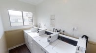 This shared laundry facility is designed for practicality and convenience, offering residents access to multiple commercial-grade washing machines and dryers arranged in a well-organized layout. The room features crisp white walls accented by crown molding near the ceiling, lending a clean and polished appearance to the utilitarian space. Dark hardwood-style flooring contrasts with the bright white appliances and walls, adding a subtle design element to the room. A window on the exterior wall allows natural light to fill the space, creating a bright and pleasant environment. Posted laundry room rules and operating hours are clearly displayed on the wall, ensuring users are well-informed. Electrical outlets are conveniently positioned along the walls to accommodate additional needs. The Space Queen commercial washers and accompanying dryers are arranged to maximize accessibility, making laundry tasks efficient and straightforward. Overall, the laundry room is a well-maintained, functional shared amenity that prioritizes cleanliness and ease of use for its occupants.