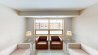 This thoughtfully designed single-floor residence offers a cohesive layout that balances functionality with a calm, neutral aesthetic throughout. The interior is finished in warm neutral tones, with light wood accents appearing consistently across cabinetry, window frames, and closet doors, lending a unified and welcoming character to the space.

The kitchen is fully equipped with stainless steel appliances, including a full-size refrigerator, gas range, dishwasher, and under-cabinet range hood. Light wood cabinetry extends from floor to ceiling, providing generous storage, while the clean countertops offer ample workspace. Tile flooring completes the practical design of this area.

The living area features a mix of seating — a tan leather sofa and armchairs paired with glass-top coffee tables — arranged to encourage conversation and relaxation. Natural light enters through wood-framed windows, offering views of the surrounding urban environment.

The bedroom is a restful retreat, furnished with a full-size bed flanked by nightstands and warm lamps. Light wood bi-fold closet doors provide practical storage, and a window allows additional natural light to filter in.

The bathroom is clean and efficiently arranged, featuring a single-sink vanity with light wood cabinetry, a stone-toned countertop, a large mirror with overhead vanity lighting, and a shower-tub combination with a curtain enclosure.