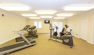 Located at 32 Blacks Ln in Springfield, NJ, this single-floor fitness center offers a well-appointed exercise space designed to support an active lifestyle. The room is finished with neutral-toned walls, crown molding, and wall-to-wall carpeting that lends a clean, comfortable atmosphere throughout.

The exercise room is generously sized, accommodating a well-rounded selection of cardio equipment including treadmills, an elliptical trainer, and both upright and recumbent stationary bikes. Multiple flush-mounted ceiling light panels ensure bright, even illumination across the entire workout area. A wall-mounted flat-screen television provides entertainment during workouts, while large windows with vertical blinds invite natural light and offer views of the surrounding greenery.

Access to the fitness area is through a set of wide double doors, making entry straightforward and accessible. A dedicated restroom is conveniently located adjacent to the exercise room, featuring tile flooring, a single-sink vanity with wood cabinetry, and a safety grab bar — reflecting a thoughtful, user-friendly design. The restroom is also equipped with a fire alarm, underscoring the facility's attention to safety. Overall, this fitness center provides a functional, tidy, and welcoming environment for daily exercise.