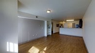 This single-floor space offers an open and functional layout highlighted by warm hardwood flooring that runs throughout, lending a cohesive and inviting atmosphere to the interior. The main living area is generously sized, featuring neutral-toned walls that provide a clean, versatile backdrop. A built-in shelving unit adds both practical storage and architectural character to the space, while multiple electrical outlets along the walls offer convenience and flexibility for various arrangements.

Transitioning seamlessly from the main living area, a raised counter bar marks the boundary of the kitchen zone, which features light-colored cabinetry and overhead fluorescent lighting that ensures excellent task visibility. The kitchen layout is efficient and well-integrated with the open floor plan, making it ideal for both everyday use and entertaining.

The space benefits from flush-mount ceiling lighting in the main area, complemented by natural light that filters in and casts warm tones across the hardwood floors. A hallway visible in the background suggests additional rooms or utility spaces beyond the primary open area. Overall, the space presents a straightforward, practical design with clean lines, ample room to move, and a comfortable, well-lit ambiance throughout.