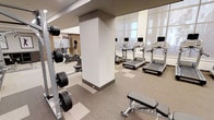 This fitness center offers a thoughtfully designed workout environment spread across a single level. The space features a contemporary aesthetic, with neutral-toned flooring in a subtle geometric pattern and crisp white walls that create a bright, clean atmosphere. Large floor-to-ceiling mirrors line one wall, visually expanding the space and allowing users to monitor their form during exercise. A row of Life Fitness treadmills is positioned along the mirrored wall, each unit equipped with its own display console for tracking workout metrics. The free weights area is well-stocked with a range of dumbbells and barbells on upright racks, accompanied by a dedicated weight bench for strength training exercises. Additional cardio equipment, including a stationary bike, is visible toward the rear of the space. Structural columns are neatly integrated into the layout without disrupting the flow of the floor plan. The overall ambiance is professional and functional, with ample spacing between equipment to ensure comfortable movement throughout. The combination of modern equipment, reflective surfaces, and a well-organized layout makes this fitness center a practical and inviting space for a full range of workout routines.