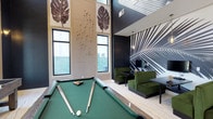 Located at 4320 Commons Dr W in Destin, Florida, this contemporary amenity lounge is a standout communal space designed with both style and function in mind. The space immediately draws the eye upward with its soaring double-height ceilings, creating an open, airy atmosphere that feels expansive and inviting. Large windows line the walls, flooding the interior with generous natural light and enhancing the sense of volume throughout.

The lounge is anchored by a full-size billiards table, positioned as a central focal point beneath the dramatic vertical wall panels adorned with sculptural tropical leaf artwork. The light-toned wood flooring grounds the space with a clean, modern aesthetic that flows seamlessly throughout.

A boldly designed accent wall featuring a large-scale black-and-white tropical palm frond mural defines the lounge seating area, where rich forest-green upholstered chairs and dark tables create an intimate gathering space ideal for socializing or working. The contrast between the dark, moody accent wall and the light, neutral tones elsewhere gives the space a dynamic, design-forward character.

Overall, the space balances a tropical coastal influence with sleek, modern finishes, making it a sophisticated and functional retreat well-suited to the vibrant Destin, Florida lifestyle.