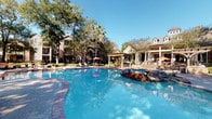 This outdoor community space centers around a generously sized, freeform resort-style swimming pool with crystal-clear blue water, a decorative rock water feature, and a gradual entry point for easy access. The pool deck is finished with attractive brick paver hardscaping and is outfitted with multiple lounge chairs and umbrella-shaded dining sets, providing ample space for both relaxation and socializing. A pergola structure adds a shaded gathering spot near the pool's edge, enhancing the resort-like atmosphere.

The surrounding grounds are thoughtfully landscaped with a variety of mature trees — including tall palms and established deciduous trees — along with manicured shrubs and well-maintained lawn areas that provide a lush, green backdrop. Wrought iron fencing defines portions of the perimeter, adding a classic aesthetic while maintaining an open feel.

The residential buildings framing the courtyard are constructed of warm red brick with stucco accents, dormer windows, multi-pane bay windows, and private balconies, reflecting a traditional architectural style. Pathway lighting and benches are integrated throughout the grounds, contributing to the space's functionality. The overall ambiance is that of a well-maintained, resort-inspired outdoor retreat designed for community enjoyment in a comfortable, park-like setting.