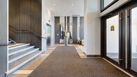 Located at 3488 Ketcheson Rd in Richmond, British Columbia, this building's lobby and hallway area presents a polished, contemporary design that makes a strong first impression. The entrance foyer is anchored by striking floor-to-ceiling marble wall cladding in soft grey and white tones, which frames a stainless steel elevator and an adjacent staircase with sleek metal railings. Large-format stone-effect floor tiles in warm taupe and grey are laid in an alternating pattern, adding visual depth to the space, while a wide carpet runner guides visitors from the entrance inward.

The lobby opens into a spacious, well-lit corridor lined with dark wood-panelled columns and walls that contrast elegantly with the lighter tile and white ceilings. Recessed pot lighting throughout ensures the area is evenly and warmly illuminated. Tall decorative planters with sculptural floral arrangements are strategically placed to soften the architectural lines and add a welcoming, natural touch.

A dedicated parcel room off the main corridor houses modern smart package lockers alongside additional secure storage units, offering a practical and organized solution for residents. The hallway extends further with the same refined tile flooring, providing access to additional building amenities. Overall, the space exudes a high-end residential building aesthetic, balancing sophisticated materials with functional design.