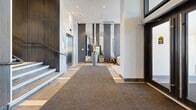Located at 3488 Ketcheson Rd in Richmond, British Columbia, this building's lobby and hallway area presents a polished, contemporary design that makes a strong first impression. The entrance foyer is anchored by striking floor-to-ceiling marble wall cladding in soft grey and white tones, which frames a stainless steel elevator and an adjacent staircase with sleek metal railings. Large-format stone-effect floor tiles in warm taupe and grey are laid in an alternating pattern, adding visual depth to the space, while a wide carpet runner guides visitors from the entrance inward.

The lobby opens into a spacious, well-lit corridor lined with dark wood-panelled columns and walls that contrast elegantly with the lighter tile and white ceilings. Recessed pot lighting throughout ensures the area is evenly and warmly illuminated. Tall decorative planters with sculptural floral arrangements are strategically placed to soften the architectural lines and add a welcoming, natural touch.

A dedicated parcel room off the main corridor houses modern smart package lockers alongside additional secure storage units, offering a practical and organized solution for residents. The hallway extends further with the same refined tile flooring, providing access to additional building amenities. Overall, the space exudes a high-end residential building aesthetic, balancing sophisticated materials with functional design.