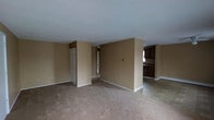 This single-floor residence offers a straightforward and functional layout designed for comfortable everyday living. The main living area is finished with wall-to-wall carpet in a warm neutral tone, complemented by beige-painted walls and white trim that create a clean, cohesive look throughout. A textured ceiling adds a classic touch to the space.

A defining feature of the living area is a partial partition wall that helps define the room without fully closing it off, maintaining an open and connected feel. A bifold closet door provides convenient built-in storage, adding to the practicality of the space.

The living area transitions seamlessly into the kitchen, which is distinguished by tile flooring, dark wood cabinetry, and a countertop workspace. The kitchen is positioned to allow natural light to filter through, brightening the adjacent areas. A baseboard heating unit runs along the perimeter, ensuring efficient climate control throughout the colder months.

The overall ambiance is simple, neutral, and move-in ready, offering a blank canvas that can be easily personalized. The open sightlines between the living and kitchen areas make the space feel connected and versatile, suitable for a range of living arrangements.