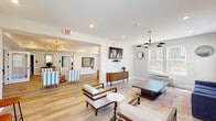 Located at 35 Violet Ave in Poughkeepsie, New York, this thoughtfully designed single-floor amenity space offers a well-rounded blend of comfort, style, and functionality. The open-concept layout flows seamlessly between multiple distinct areas, all unified by light hardwood flooring, neutral wall tones, and abundant natural light streaming through large windows.

The main lounge area serves as the heart of the space, furnished with mid-century modern accent chairs in blue-and-white stripes, a bold navy sofa, wooden armchairs, and a geometric coffee table. Statement sputnik-style brass chandeliers add a warm, contemporary touch overhead. A wall-mounted flat-screen TV and a wooden credenza complete the relaxed gathering atmosphere. French doors open to an outdoor view, further brightening the space.

An adjacent seating nook offers additional lounge seating centered around a textured woven ottoman, with a decorative chess board mounted on the wall providing a playful, social element.

The kitchenette features white shaker-style cabinetry with matte black hardware, granite countertops, a stainless steel sink, and elegant gold wall sconces — ideal for casual refreshments.

The fitness center is comprehensively equipped with treadmills, an elliptical machine, a weight bench, dumbbells, medicine balls, and a wall-mounted television, offering residents a convenient and fully functional workout environment.