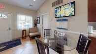 This single-floor space offers an open, well-connected layout highlighted by vaulted ceilings with recessed lighting that enhance the sense of volume and airiness throughout. The flooring transitions between large-format tile in a warm travertine tone and white ceramic tile, lending visual continuity across the different functional areas.

The main living and reception area is spacious and inviting, featuring vaulted ceilings, large windows with vertical blinds that allow generous natural light, and seating arrangements that include a cushioned bench and a glass-top dining table with dark wood chairs. A wall-mounted flat-screen television and framed artwork add character to the space, while a gas fireplace framed by a decorative mirror serves as a focal point, creating a warm and welcoming ambiance.

Adjacent to the main area, the kitchen is efficiently designed with rich dark wood cabinetry, light countertops, and stainless steel appliances including a refrigerator, electric range, and microwave. A double-basin sink and ample cabinet storage above and below the counters provide both functionality and visual appeal.

Multiple interior doors lead to additional rooms, and a paneled entry door with a transom window introduces natural light at the entrance. Overall, the space presents a comfortable and functional environment with a warm, neutral color palette throughout.