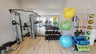 This modern fitness center at 1100 E Osborn Rd in Phoenix, Arizona, is a well-appointed, single-floor facility designed to support a wide range of workout styles and fitness goals. The space features light wood-look flooring, neutral gray walls, and recessed lighting throughout, creating a clean and energizing environment.

The cardio zone is anchored by a row of True-brand treadmills and elliptical cross-trainers positioned along large windows that overlook lush greenery and the outdoor pool area, allowing ample natural light to fill the space. A wall-mounted flat-screen TV provides entertainment during workouts.

The strength training area is equipped with a dual-cable functional trainer and a dedicated weight bench, ideal for a full range of resistance exercises. A tiered dumbbell rack holds an extensive selection of free weights to accommodate all fitness levels. Floor-to-ceiling mirrors line one wall, enhancing the sense of space and allowing users to monitor their form.

A functional and flexibility zone rounds out the facility, featuring a multi-ball storage rack stocked with stability balls in assorted sizes, resistance bands, yoga mats, and a stationary bike. A leg extension/curl machine provides additional targeted strength training options.

The space offers direct access to the outdoors through a glass door, further connecting the interior to the surrounding environment. Overall, this fitness center is thoughtfully organized, well-lit, and comprehensively equipped.