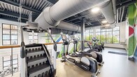This well-appointed two-story fitness center offers an extensive and thoughtfully designed space for a comprehensive workout experience. The main floor provides an expansive area housing a wide array of strength training equipment, free weights, and cable machines, all set against large floor-to-ceiling windows that overlook an outdoor pool and the surrounding community. The open layout is enhanced by polished flooring, full-length mirrors, and bright, recessed lighting that creates an energizing atmosphere.

The upper level features a dedicated cardio zone equipped with treadmills, ellipticals, and stationary bikes, all positioned along generous windows that invite abundant natural light and outdoor views. Exposed ductwork and an industrial-style open ceiling add a contemporary edge, while a striking oversized fan anchors the double-height space connecting both levels.

Separate studio rooms serve varied fitness needs — one features warm hardwood flooring and a vibrant, full-wall mural for group or solo training sessions, while another is outfitted with a heavy bag, rowing machine, functional training rig, and a wall-mounted display screen for more intensive workouts.

An outdoor terrace rounds out the facility, offering open-air seating with sweeping views of the pool area and the rolling hills of the surrounding neighborhood — an ideal space to cool down or relax after a workout.