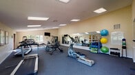 This single-floor fitness center offers a comprehensive workout environment designed to support a wide range of exercise routines. The main workout area is generously proportioned, featuring high vaulted ceilings that lend an open, airy feel to the space. The room is finished with durable, wall-to-wall blue carpet and illuminated by a combination of recessed and flush-mounted ceiling light panels that ensure bright, even lighting throughout.

The facility is well-appointed with a variety of cardio equipment, including treadmills, an elliptical trainer, a rowing machine, and a stationary bike, all arranged to allow comfortable use. A dedicated strength and flexibility zone lines one wall, featuring a multi-station cable machine, a rack of colorful medicine balls and exercise balls in various sizes, resistance bands, and yoga mats. Full-length wall mirrors run along multiple walls, enhancing the sense of space and allowing users to monitor their form during workouts. A wall-mounted television adds entertainment for cardio sessions.

An adjoining entry and transition area features tile flooring, warm neutral walls, and large windows on multiple sides that provide views of adjacent outdoor tennis courts and lush greenery, filling the space with natural light. Direct exterior access through glass-paneled doors connects the fitness center to the surrounding outdoor amenities, creating a seamless indoor-outdoor experience.