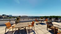 This space features a standout rooftop terrace that serves as its centerpiece, offering expansive, panoramic views of the surrounding cityscape and horizon under open skies. The terrace is finished with large-format tile flooring and is equipped with multiple outdoor dining sets, including wooden tables and chairs arranged to take full advantage of the sweeping vistas. Decorative planters with colorful flowers add a touch of greenery and vibrancy to the outdoor area, enhancing its welcoming ambiance. The rooftop's low-profile perimeter railing ensures unobstructed sightlines across the urban landscape, making it well-suited for both relaxed gatherings and al fresco dining.

Complementing the rooftop is a bathroom finished in warm, neutral tones. The bathroom features a classic wood-trimmed vanity with a white countertop and integrated sink, chrome faucet hardware, and a framed rectangular mirror above. Warm-toned walls create a cozy atmosphere, and the space is outfitted with practical details including a wall-mounted towel ring and a small decorative shelf above the toilet. Brass-toned hardware and switch plates throughout add a consistent, traditional finish to the interior details, tying the bathroom's overall aesthetic together cohesively.