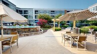 Located at 101 Bryce Ln in King of Prussia, PA, this thoughtfully designed outdoor courtyard offers a resort-inspired retreat set within a modern multi-story building complex. The space spans two levels and is anchored by a generously sized swimming pool with a dedicated wading area, both framed by a wooden pool deck lined with lounge chairs and market umbrellas. Adjacent to the pool, a stone-accented hot tub or wading feature adds a refined touch, complemented by a gated entry and accessible ramp.

The surrounding landscape is meticulously maintained, featuring a central open lawn area bordered by curved mulched garden beds, mature shade trees, ornamental grasses, flowering shrubs, and seasonal plantings. Winding concrete pathways and railed walkways connect the various zones throughout the courtyard, creating an accessible and cohesive flow. A pergola-covered outdoor dining area with wooden tables and chairs provides a comfortable space for al fresco gatherings, while Adirondack chairs and benches are scattered throughout for casual relaxation.

The building facades enclosing the courtyard feature a contemporary architectural palette of gray siding, large windows, and private balconies on upper floors. A glass-fronted amenity room, likely housing fitness or clubhouse facilities, opens directly onto the pool area, enhancing the indoor-outdoor connection. The entire space benefits from abundant natural light and a sense of verdant, park-like seclusion.