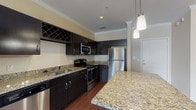 This single-floor, one-bedroom, one-bathroom residence combines thoughtful design with quality finishes across its well-proportioned layout. The kitchen serves as a standout feature, equipped with stainless steel appliances including a refrigerator, range, and built-in microwave. Rich dark cabinetry contrasts elegantly with speckled granite countertops, while a subway tile backsplash and an integrated wine rack add refined detail. A granite-topped island with pendant lighting above creates a natural divide between the kitchen and the open living area, which features warm hardwood flooring and crown molding throughout. Natural light pours into the living space through a bank of windows fitted with blinds, and a ceiling fan enhances comfort. The bedroom continues the warm, neutral palette with plush carpeting, a ceiling fan, and a window that invites natural light. A connecting hallway leads to the full bathroom, which mirrors the kitchen's design aesthetic with matching dark cabinetry, granite countertops, and a large framed mirror above the vanity. Dark hardwood flooring in the wet areas ties the space together cohesively, creating a consistent and polished look throughout the entire residence.