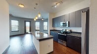 This single-floor unit showcases a contemporary design with a cohesive, open layout connecting the kitchen and living areas. The kitchen serves as the focal point, featuring a central island with granite countertops and an undermount sink, complemented by dark cabinetry, a subway tile backsplash, and a full suite of stainless steel appliances including an electric range, over-the-range microwave, and bottom-freezer refrigerator. Pendant lighting above the island and track lighting overhead enhance both functionality and ambiance.

Rich hardwood-style flooring extends throughout the main living areas, adding warmth to the modern palette of gray and neutral tones. The living area is distinguished by a striking curved wall lined with multiple windows, allowing generous natural light and outdoor views, while a ceiling fan provides additional comfort. A secondary bedroom features neutral carpeting, dual windows, and a ceiling fan.

Both bathrooms are finished consistently, each offering granite-topped dark vanities, undermount sinks, large-format gray tile surrounding the soaking tubs, and dark cabinetry for storage. The overall design maintains a clean, urban aesthetic throughout, unified by consistent fixtures, finishes, and attention to detail.