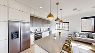 This contemporary single-floor residence showcases a clean, modern design aesthetic defined by a neutral palette, light hardwood flooring, and abundant natural light throughout. The open-concept kitchen is a standout feature, boasting two-tone cabinetry — crisp white upper cabinets paired with warm wood-toned lower units — complemented by a marble-style backsplash, sleek grey quartz countertops, and stainless steel appliances. A large central island with seating for multiple guests serves as both a functional workspace and a social hub, illuminated by a trio of striking brushed gold pendant lights. The living area is bright and airy, with floor-to-ceiling black-framed windows and a sliding glass door that opens to an outdoor balcony, offering views of the surrounding cityscape and greenery. The bedroom is a tranquil retreat, featuring expansive windows with tree-lined outdoor views, a wall-mounted television, and generous natural light. Two bathrooms maintain the home's modern sensibility — one features a glass-enclosed walk-in shower with white subway tile, black accents, and brushed gold hardware, while the second offers a clean, minimalist layout with dark slate-style tile flooring and white cabinetry. Together, these spaces form a cohesive, well-appointed residence with refined urban style.