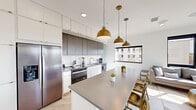 This contemporary single-floor residence showcases a clean, modern design aesthetic defined by a neutral palette, light hardwood flooring, and abundant natural light throughout. The open-concept kitchen is a standout feature, boasting two-tone cabinetry — crisp white upper cabinets paired with warm wood-toned lower units — complemented by a marble-style backsplash, sleek grey quartz countertops, and stainless steel appliances. A large central island with seating for multiple guests serves as both a functional workspace and a social hub, illuminated by a trio of striking brushed gold pendant lights. The living area is bright and airy, with floor-to-ceiling black-framed windows and a sliding glass door that opens to an outdoor balcony, offering views of the surrounding cityscape and greenery. The bedroom is a tranquil retreat, featuring expansive windows with tree-lined outdoor views, a wall-mounted television, and generous natural light. Two bathrooms maintain the home's modern sensibility — one features a glass-enclosed walk-in shower with white subway tile, black accents, and brushed gold hardware, while the second offers a clean, minimalist layout with dark slate-style tile flooring and white cabinetry. Together, these spaces form a cohesive, well-appointed residence with refined urban style.