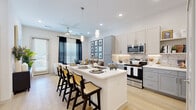 This efficiently designed one-bedroom, one-bathroom unit offers a thoughtfully organized layout across a single floor. The open-concept kitchen and living area serves as the heart of the unit, featuring light wood-style flooring, recessed lighting, and a ceiling fan that enhance the bright, airy atmosphere. The kitchen is well-appointed with two-tone grey and cream shaker-style cabinetry, stainless steel appliances including a refrigerator, microwave, and range, a patterned tile backsplash, and a central island with a built-in sink and seating for multiple bar stools. Pendant lights above the island add a modern touch. The adjacent living area benefits from natural light streaming through large windows dressed with blinds and dark curtain panels, with access to an exterior door. The bedroom is a comfortable retreat, offering a window with natural light, a dedicated closet, and neutral tones throughout. The bathroom features large-format tile flooring, marble-look tile surrounding a soaking tub and shower combination, built-in open shelving for storage, and a vanity with ample cabinet space.