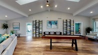 Located at 6550 150th Ave N in Largo, Florida, this single-story community clubhouse offers a well-rounded amenity space designed for recreation, fitness, and productivity. The main lounge area is a welcoming gathering space anchored by a striking stone-accent fireplace wall with a mounted flat-screen TV. Warm hardwood-style flooring, teal and neutral coastal-inspired furnishings, abundant natural light from large windows, and vaulted ceilings with exposed beams and skylights create an open, airy atmosphere throughout. An adjacent game room features soaring vaulted ceilings, decorative ladder shelving units, a shuffleboard table, and additional seating, making it ideal for social gatherings. The fitness center is spacious and well-equipped with multiple treadmills, elliptical machines, a multi-station weight machine, and a free-weight rack, all set against bold teal accent walls with ample natural light. A separate business and work area provides a built-in desk with dark cabinetry, counter seating, and bistro-height tables, offering a quiet space for focused work or meetings. Together, these amenity areas deliver a comprehensive and thoughtfully designed community facility.