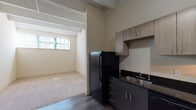 Located in the heart of downtown Winston-Salem, this loft-style unit at 231 W Fifth St blends industrial character with modern updates. The space is defined by its striking exposed structural beams and ductwork overhead, lending an authentic urban aesthetic throughout.

The kitchen is well-appointed and fully equipped, featuring black appliances including a refrigerator, electric range, and dishwasher. Dark granite-style countertops contrast with light wood-grain cabinetry that extends to the upper walls, providing ample storage. A pendant light fixture adds a refined touch above the kitchen area, which transitions seamlessly onto durable wood-style flooring.

The living area is generously sized, finished with plush neutral carpet that adds warmth and comfort underfoot. A horizontal window along the lower portion of the wall allows natural light to filter in, illuminating the open, airy feel of the room. The exposed beams and pipes above maintain the industrial charm that defines the unit's character.

The bathroom is clean and modern, featuring a dark-toned vanity with an undermount sink, dark countertop, a large mirror with overhead lighting, and a walk-in shower stall. The overall design balances functionality with a contemporary aesthetic, making this a practical and distinctive space in a prime Winston-Salem location.