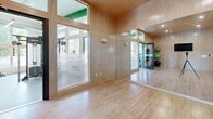 Located at 700 Bloomfield Ave in Bloomfield, Connecticut, this single-floor fitness facility offers a generously sized, well-appointed exercise space designed to accommodate a wide range of workout needs. The main gym area features high ceilings with bold green structural beams and exposed ductwork, lending the space an open, industrial character. Large windows line the walls, flooding the floor with natural light and offering views of the surrounding greenery. The durable speckled rubber flooring throughout the main area supports a comprehensive array of equipment, including strength machines, free weights with dedicated rack storage, rowing machines, cable systems, and cardio equipment. Distinctive green-painted support columns punctuate the floor plan, reinforcing the facility's modern aesthetic. A separate studio room is enclosed with warm wood-paneled walls, a hardwood floor, frosted glass interior windows, and full-length mirrors, making it well-suited for yoga, stretching, or group fitness classes. Exercise mats are neatly stored on wall-mounted racks within the studio. Linear pendant lighting and recessed ceiling fixtures ensure consistent illumination throughout both areas. The overall design balances functionality with a clean, contemporary aesthetic, creating an inviting and professional environment for fitness.