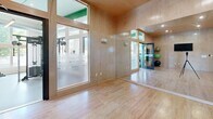 Located at 700 Bloomfield Ave in Bloomfield, Connecticut, this single-floor fitness facility offers a generously sized, well-appointed exercise space designed to accommodate a wide range of workout needs. The main gym area features high ceilings with bold green structural beams and exposed ductwork, lending the space an open, industrial character. Large windows line the walls, flooding the floor with natural light and offering views of the surrounding greenery. The durable speckled rubber flooring throughout the main area supports a comprehensive array of equipment, including strength machines, free weights with dedicated rack storage, rowing machines, cable systems, and cardio equipment. Distinctive green-painted support columns punctuate the floor plan, reinforcing the facility's modern aesthetic. A separate studio room is enclosed with warm wood-paneled walls, a hardwood floor, frosted glass interior windows, and full-length mirrors, making it well-suited for yoga, stretching, or group fitness classes. Exercise mats are neatly stored on wall-mounted racks within the studio. Linear pendant lighting and recessed ceiling fixtures ensure consistent illumination throughout both areas. The overall design balances functionality with a clean, contemporary aesthetic, creating an inviting and professional environment for fitness.