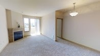 This 2-bedroom, 2-bathroom residence at Copperbend is designed with a practical and comfortable layout spread across a single floor. The main living area is anchored by plush wall-to-wall carpeting and neutral-toned walls, creating a bright and welcoming atmosphere. A corner fireplace adds a cozy focal point, while sliding glass doors lead to a private outdoor space, allowing natural light to fill the room and offering a view of the surrounding landscape. An elegant pendant light fixture adds a refined touch to the transitional space between the living area and the rest of the home. Interior doors with wood-trimmed frames connect to additional rooms, maintaining a cohesive design aesthetic throughout. The open feel of the living space, combined with ample natural light, contributes to an airy and functional environment. The neutral color palette and clean finishes throughout provide a versatile backdrop suited to a variety of interior styles, making this residence a comfortable and move-in-ready home at Copperbend.