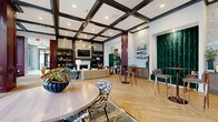 Located at 370 Carmelina Ave in Columbus, Ohio, this elegantly designed single-floor communal space serves as a grand lobby and living area for its community. The space makes an immediate impression with its striking use of deep burgundy paneling, which frames the interiors and adds a bold, refined character throughout.

The welcoming entryway features large floor-to-ceiling windows that flood the space with natural light and offer views of the outdoor courtyard and surrounding neighborhood. Light-toned herringbone-patterned flooring flows seamlessly from the entry into the main living area, providing warmth and visual continuity.

A front reception desk anchors the lobby area, accompanied by comfortable seating that invites guests to relax. Further into the space, the living room showcases a dramatic coffered ceiling with dark trim, a decorative chandelier, and a wood-burning fireplace flanked by built-in bookshelves. Curated furnishings, including a channeled sofa, eclectic accent chairs, and layered rugs, create an inviting and stylish atmosphere.

A dedicated lounge and café-style bar area with shelving and cabinetry provides additional functionality. Tall burgundy columns, decorative sconces, and abundant greenery complete the upscale aesthetic, making this space both visually stunning and highly functional for residents and visitors alike.