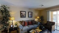 This single-floor living space centers around a generously sized gray upholstered sofa adorned with an assortment of accent pillows in rich burgundy and brown tones, creating a warm and inviting focal point. The living area is finished with smooth walls painted in a soft, neutral tone that complements the overall color palette. Two framed art prints depicting musical themes — a piano staircase and a guitar — are displayed prominently on the main wall, adding a refined, artistic character to the room. A floor lamp on the left and a table lamp on the right provide warm ambient lighting that enhances the cozy atmosphere. A large indoor tropical plant in the corner introduces a natural, organic element to the space. The coffee table in the foreground holds a collection of magazines, adding a lived-in, casual feel. A side table with a small potted plant and books anchors the left side of the room. A dark wood console or media unit is partially visible to the right, offering additional storage. The space overall presents a comfortable and thoughtfully arranged living environment with a consistent warm, earthy aesthetic throughout.