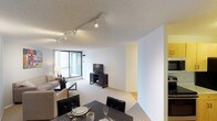 This single-floor residence features an open-concept layout that seamlessly connects the dining and living areas, creating a cohesive and functional living environment. The dining area is positioned in the foreground and is furnished with a dark-toned table and black upholstered chairs, complemented by place settings that add a polished touch. The living area flows naturally from the dining space, featuring plush carpeting, a comfortable sofa, and a media console with a flat-screen television. A floor lamp provides warm ambient lighting alongside the installed track lighting system on the textured ceiling, which allows for flexible and directional illumination throughout the space. Floor-to-ceiling windows along one wall flood the interior with abundant natural light while offering expansive city views, enhancing the sense of openness. The walls are finished in a clean white with subtle yellow accents, contributing to the bright and airy atmosphere. A transitional area visible toward the back hints at additional spaces within the unit, while the combination of carpet and hard flooring adds visual variety and practical zoning to the layout.