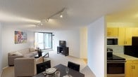 This single-floor residence features an open-concept layout that seamlessly connects the dining and living areas, creating a cohesive and functional living environment. The dining area is positioned in the foreground and is furnished with a dark-toned table and black upholstered chairs, complemented by place settings that add a polished touch. The living area flows naturally from the dining space, featuring plush carpeting, a comfortable sofa, and a media console with a flat-screen television. A floor lamp provides warm ambient lighting alongside the installed track lighting system on the textured ceiling, which allows for flexible and directional illumination throughout the space. Floor-to-ceiling windows along one wall flood the interior with abundant natural light while offering expansive city views, enhancing the sense of openness. The walls are finished in a clean white with subtle yellow accents, contributing to the bright and airy atmosphere. A transitional area visible toward the back hints at additional spaces within the unit, while the combination of carpet and hard flooring adds visual variety and practical zoning to the layout.
